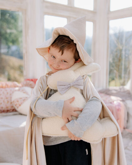 Chłopiec w pelerynie i kapeluszu czarodzieja obejmuje pluszowego królika z kokardką w jasnym pomieszczeniu