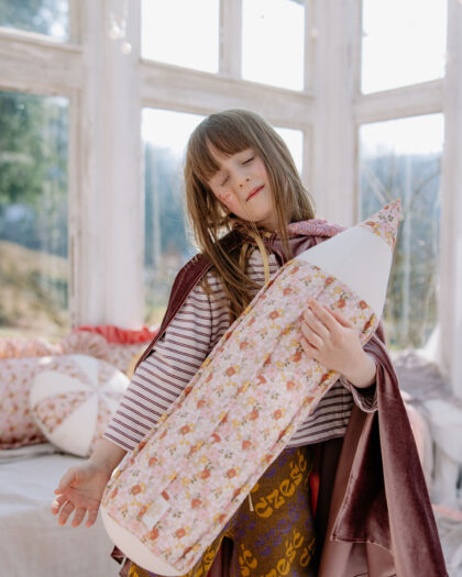 Dziewczynka w pelerynie tuląca podłużną poduszkę w kwiaty, stojąca w jasnym pokoju z dużymi oknami