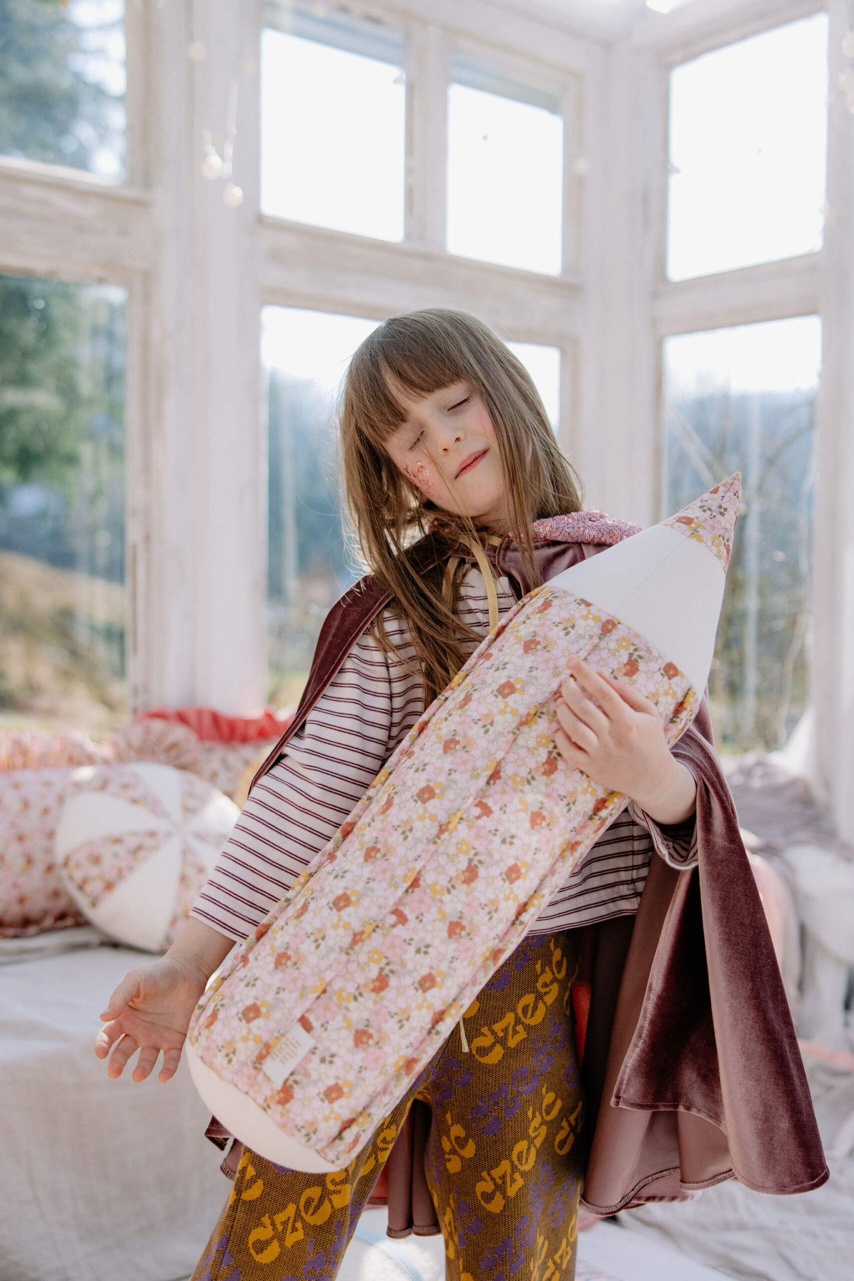 Dziewczynka w pelerynie tuląca podłużną poduszkę w kwiaty, stojąca w jasnym pokoju z dużymi oknami