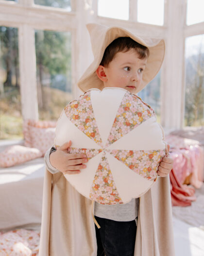 Chłopiec w kapeluszu i pelerynie trzyma ozdobną poduszkę w kwiatowe wzory, stoi przy dużych oknach