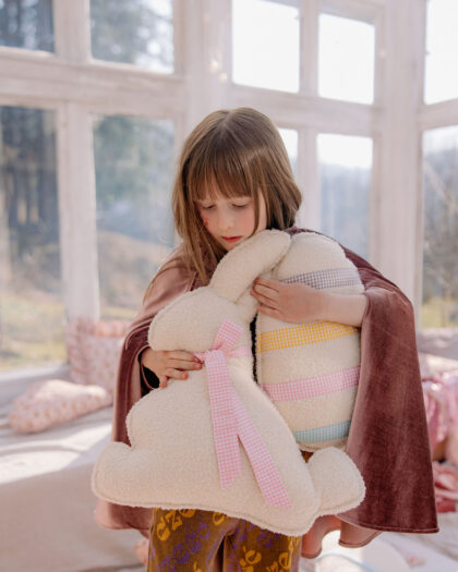 Dziewczynka obejmuje pluszowego króliczka i poduszkę w kształcie jajka w słonecznym pokoju.