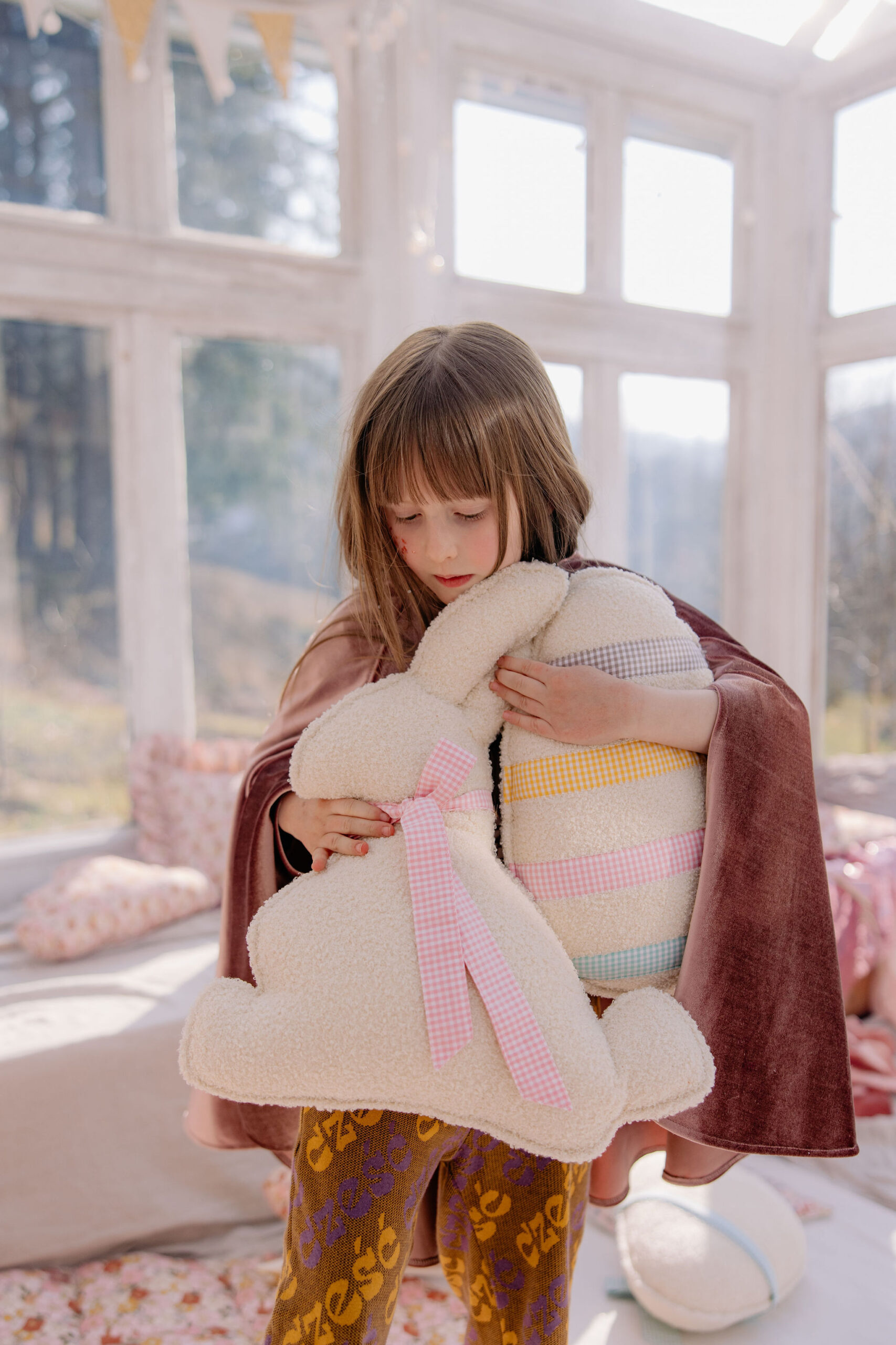 Dziewczynka obejmuje pluszowego króliczka i poduszkę w kształcie jajka w słonecznym pokoju.