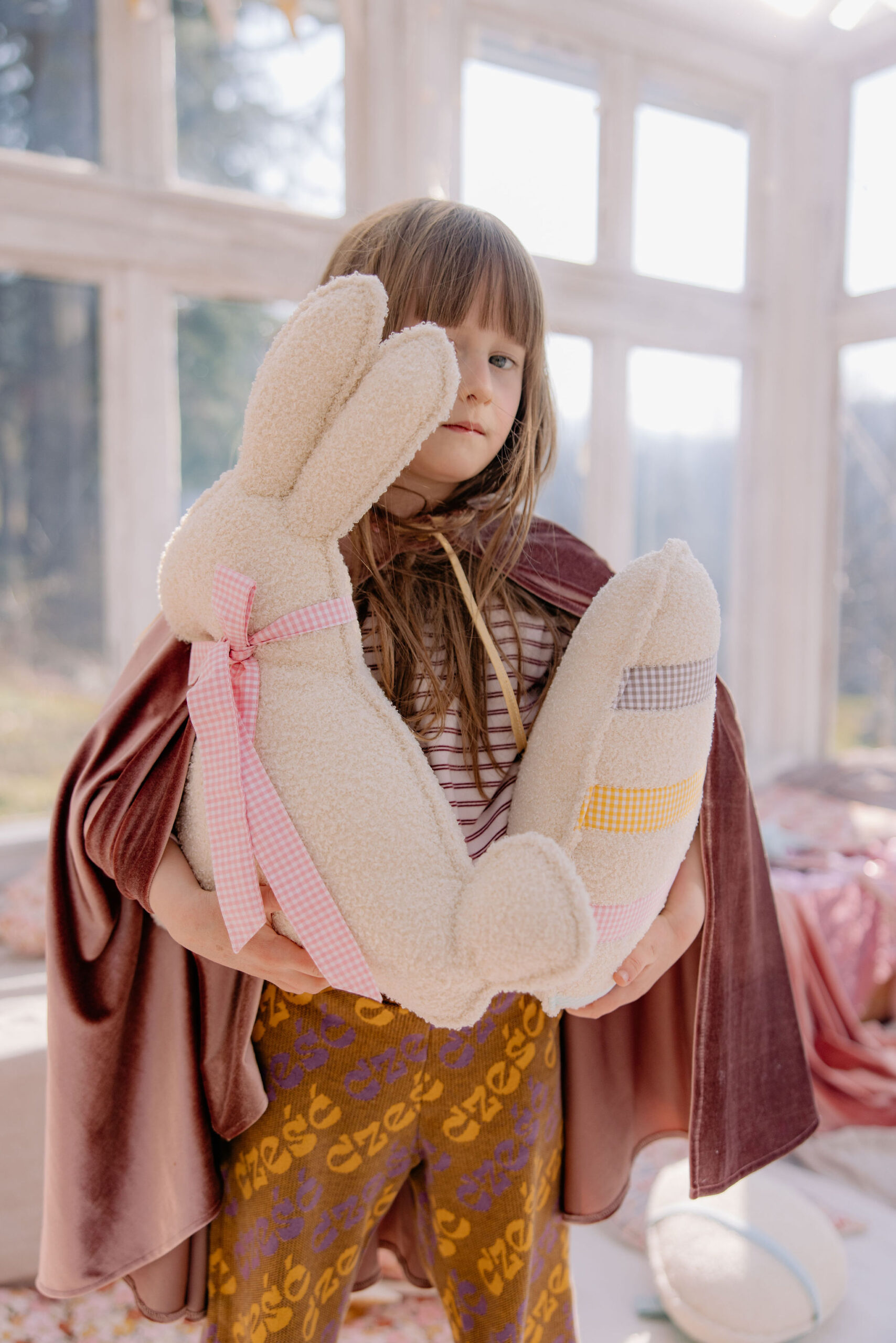Dziewczynka w pelerynie trzyma dużą miękką zabawkę królika z kokardą i kolorowymi paskami.