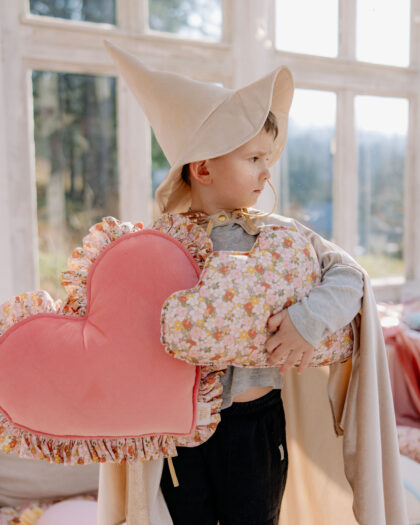 Chłopiec w kapeluszu czarodzieja trzyma dwa kwiatowe i różowe poduszki w kształcie serca przy oknie