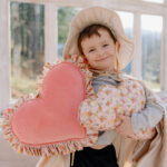 Uśmiechnięte dziecko w kapeluszu i pelerynie trzyma dwa poduszki w kształcie serca w jasnym pokoju