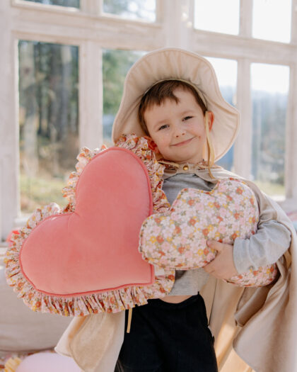 Uśmiechnięte dziecko w kapeluszu i pelerynie trzyma dwa poduszki w kształcie serca w jasnym pokoju