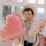 Chłopiec w kapeluszu trzyma dwa poduszki w kształcie serca, stoi w jasnym pomieszczeniu przed oknami