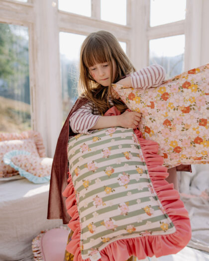 Dziewczynka z jasnobrązowymi włosami trzyma kolorowe poduszki w jasnym, słonecznym pokoju.