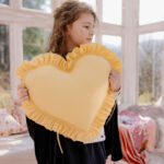 Dziewczynka trzyma żółtą poduszkę w kształcie serca z falbanką w jasnym pokoju z dużymi oknami