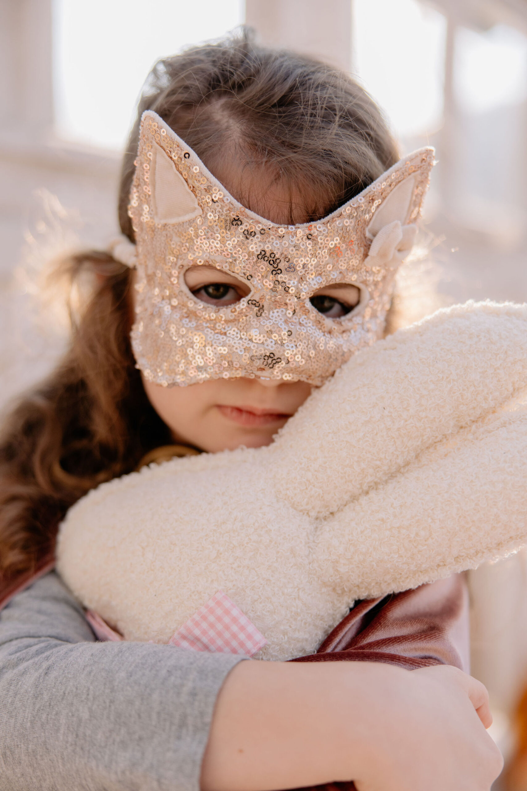 Dziewczynka w cekinowej masce kota obejmuje miękką zabawkę w jasnym pokoju oświetlonym naturalnym światłem