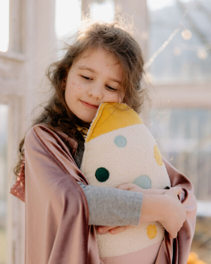 Uśmiechnięta dziewczynka przytula miękką poduszkę w kolorowe kropki, stojąc przy oknie