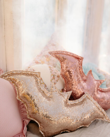 Poduszki w kształcie nietoperzy pokryte złotymi i różowozłotymi cekinami na jasnej kanapie przy oknie