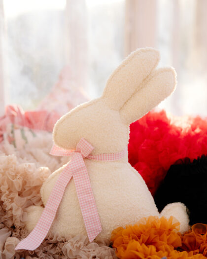 Pluszowy biały królik z różową kokardą siedzący na kolorowych falbankach w ciepłym świetle dziennym