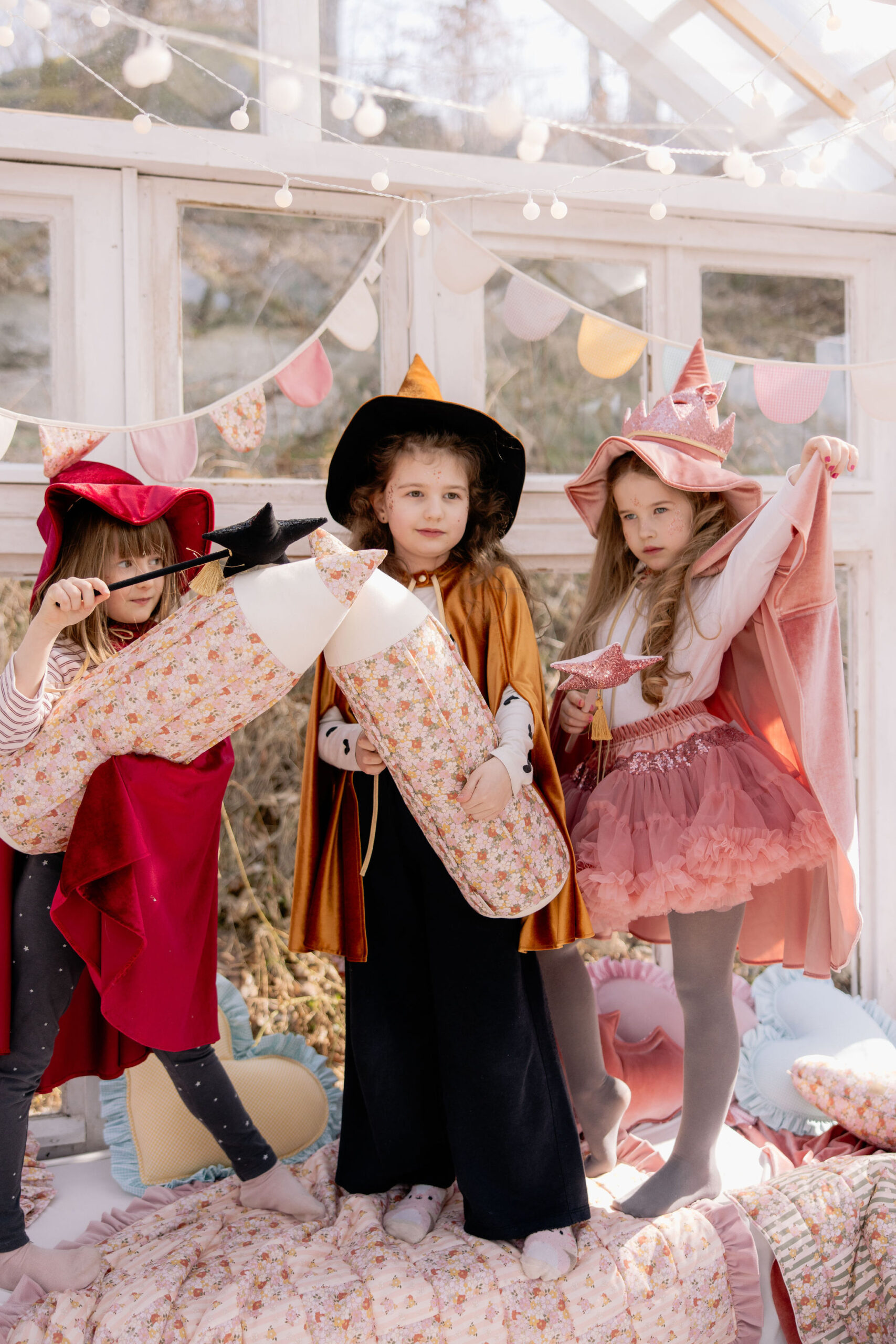 Trzy dziewczynki w kostiumach wróżek i czarownic bawiące się z poduszkami i różdżkami w jasnym pokoju