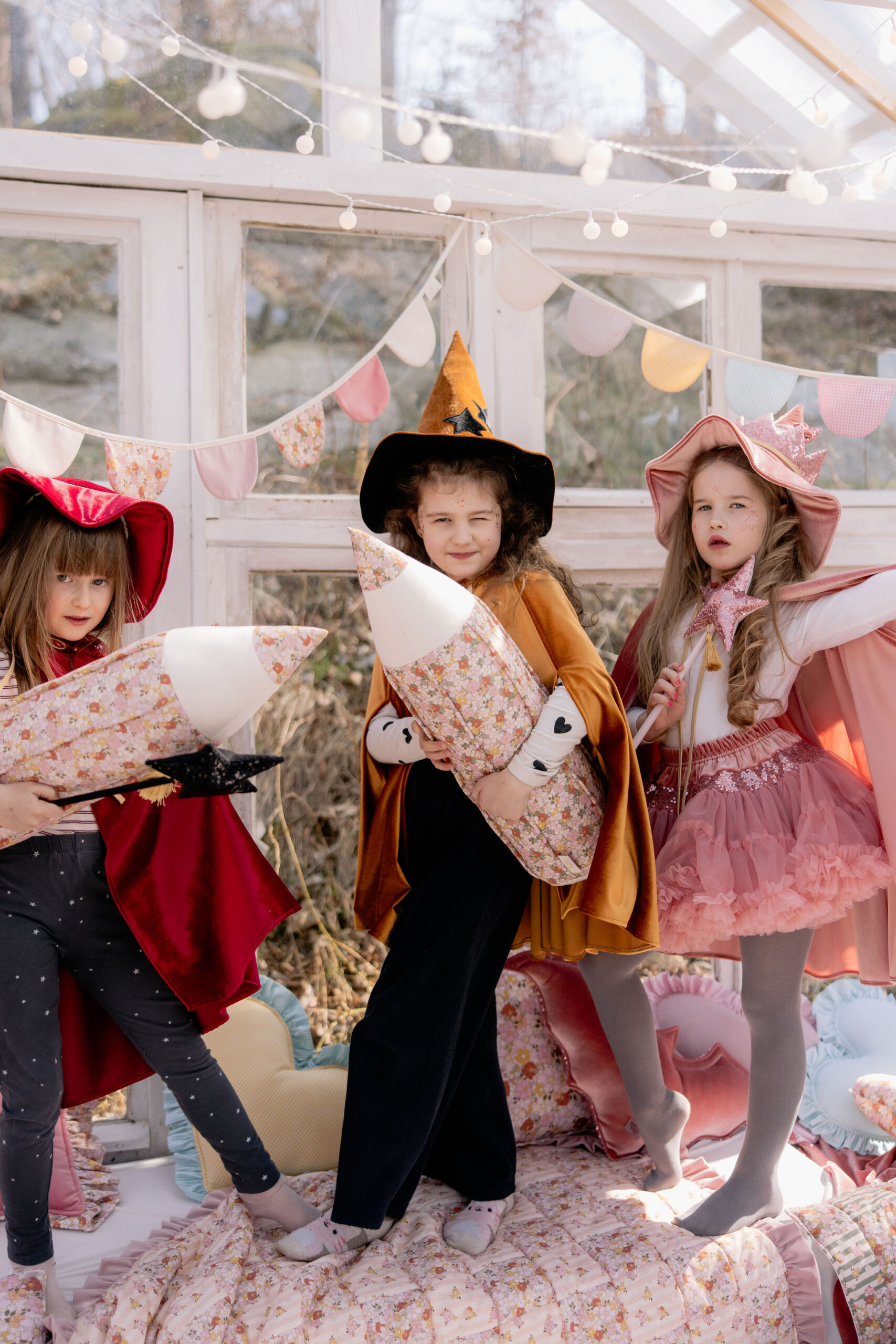 Trzy dziewczynki w kolorowych strojach czarodziejek bawią się na łóżku udekorowanym girlandami