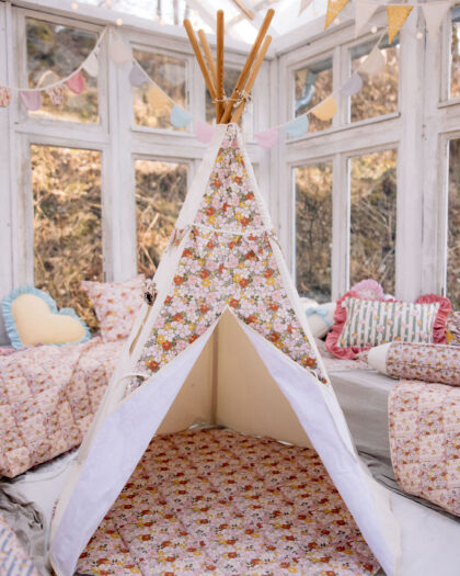 Dziecięce tipi namiotowe w jasnych kolorach z kwiatowym wzorem, otoczone poduszkami i girlandami na tle okien.
