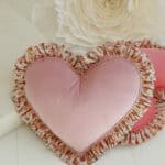 Różowa poduszka w kształcie serca z falbaną we wzór kwiatowy, na tle jasnej powierzchni i dużego białego kwiatu.