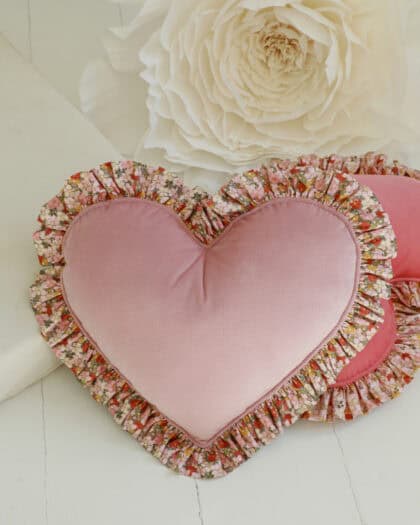 Różowa poduszka w kształcie serca z falbaną we wzór kwiatowy, na tle jasnej powierzchni i dużego białego kwiatu.