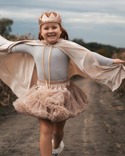 Uśmiechnięta dziewczynka w koronie i tiulowej spódniczce biega po wiejskiej drodze na tle pochmurnego nieba