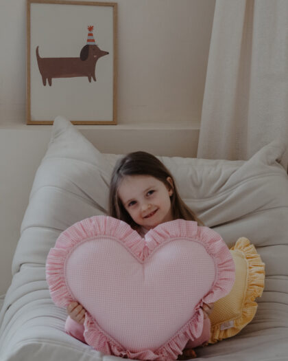 Uśmiechająca się dziewczynka trzymająca różową poduszkę w kształcie serca siedzi na białym fotelu w przytulnym pokoju