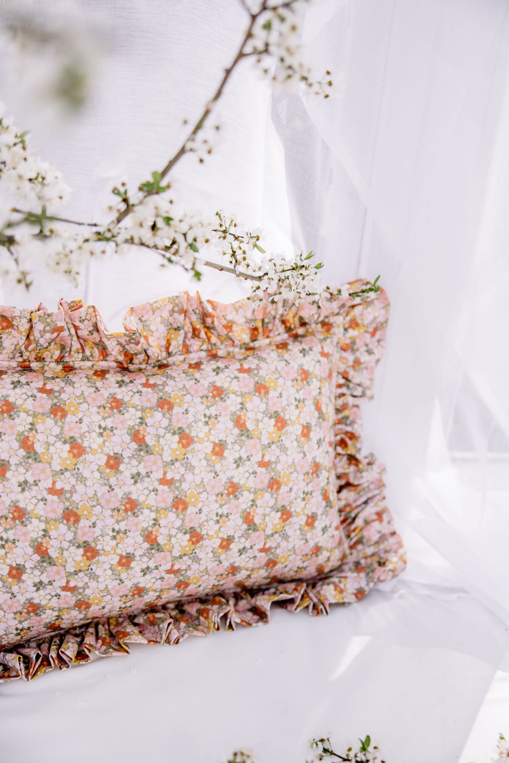 Poduszka w kwiecisty wzór z falbanką na tle białych zasłon i delikatnych gałązek kwiatów
