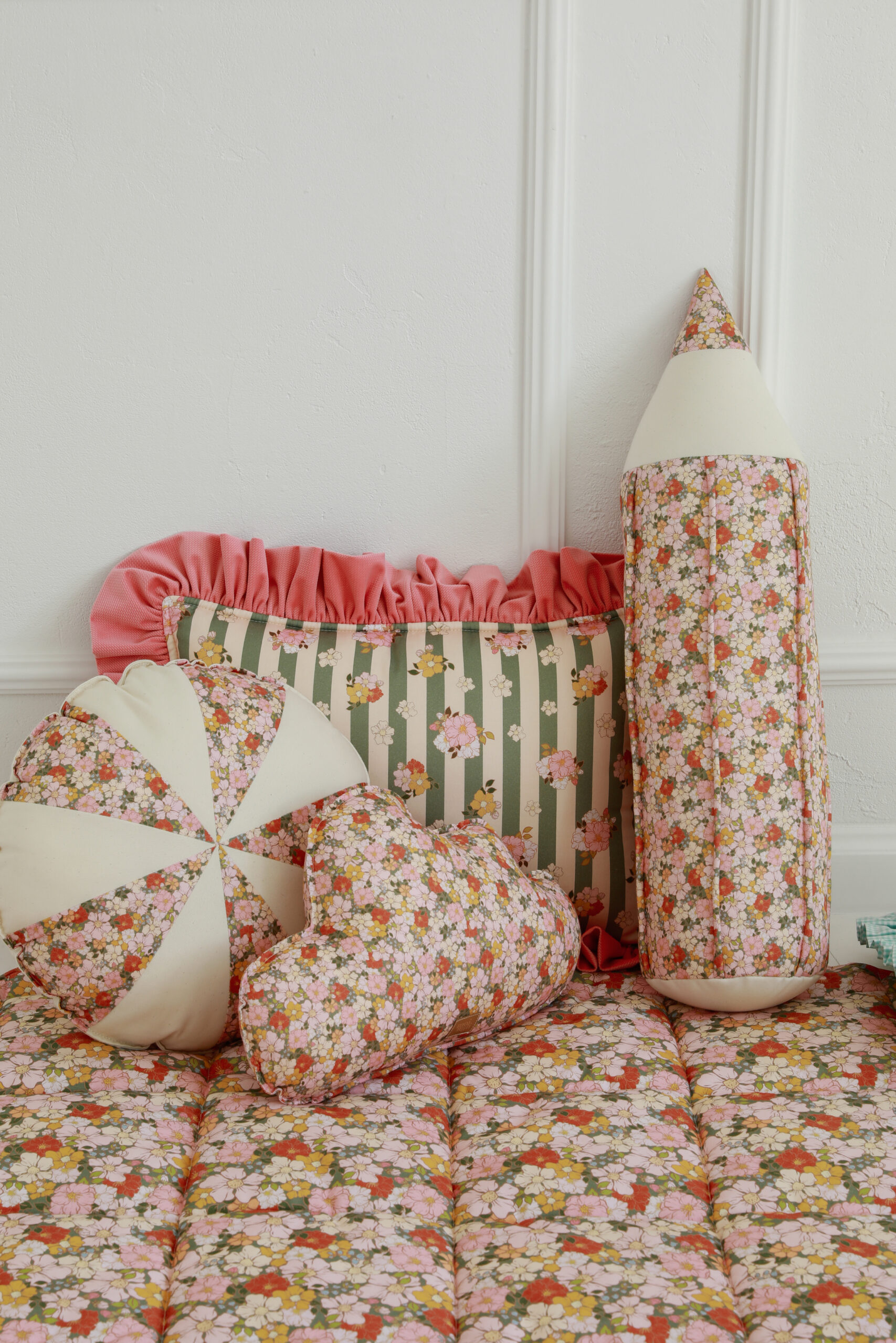 Większe i mniejsze poduszki w kwiatowe wzory na pikowanym kocyku, na tle białej ściany z panelami