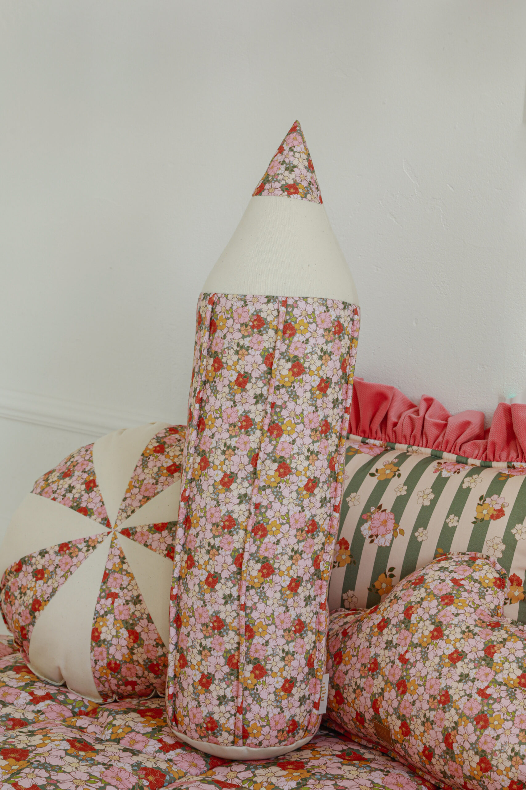 Miękka poduszka w kształcie ołówka w kwiatowy wzór na tle innych wzorzystych poduszek i pościeli.