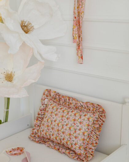 Różowo-pomarańczowa poduszka w kwiatowy wzór z falbanką na białym tle obok dużych białych dekoracyjnych kwiatów