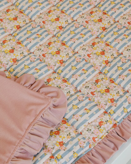 Pikowana narzuta z kwiatowym i niebiesko-białym paskowanym wzorem oraz różowym falbankowym wykończeniem.