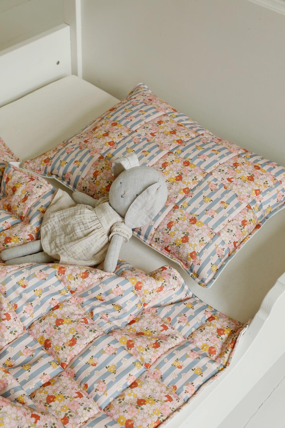 Bawełniana pościel w pastelowe kwiaty i paski oraz pluszowa szara zabawka na łóżku dziecięcym