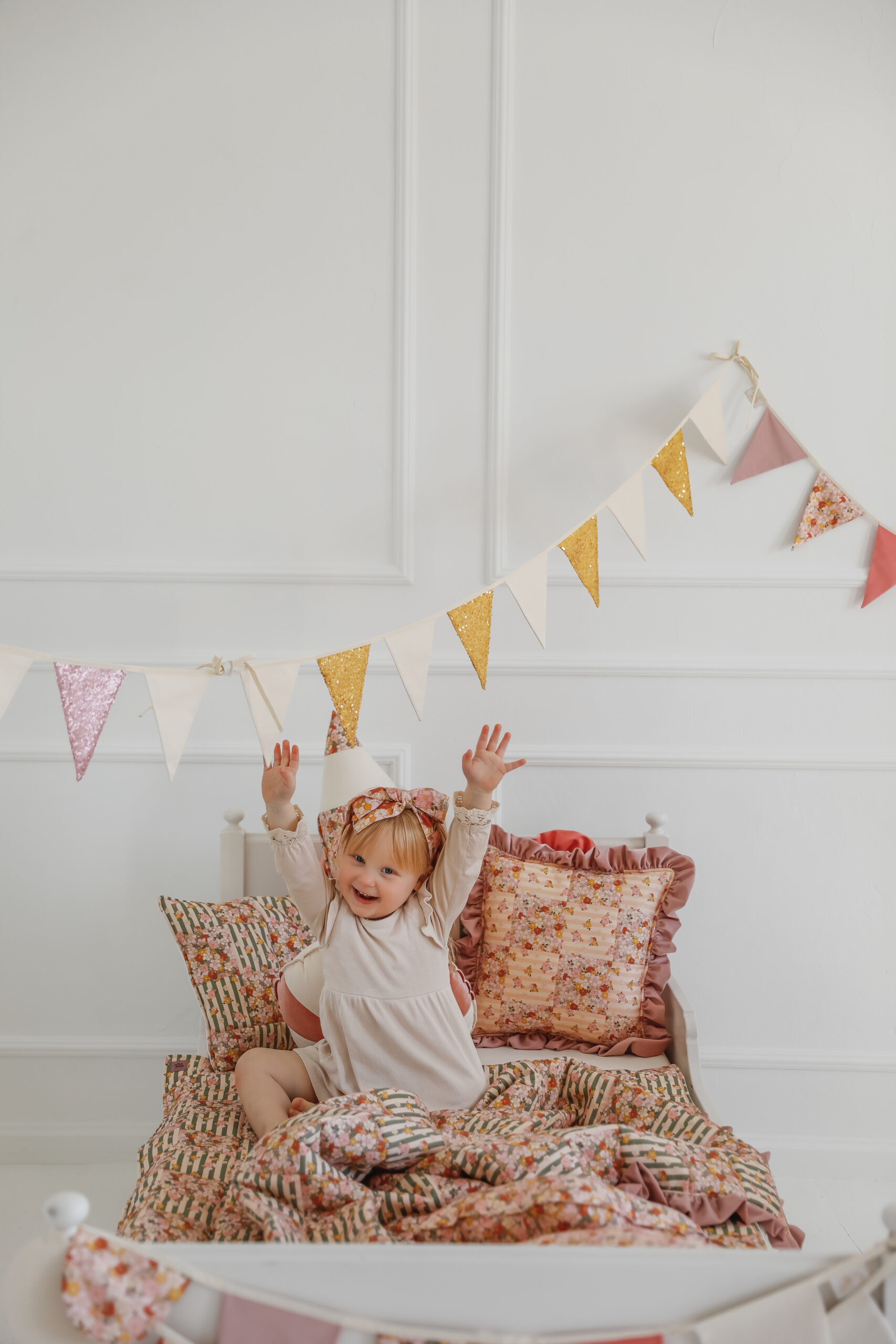 Mała dziewczynka w czapce imprezowej na łóżku z kwiatową pościelą, unosząca ręce w górę