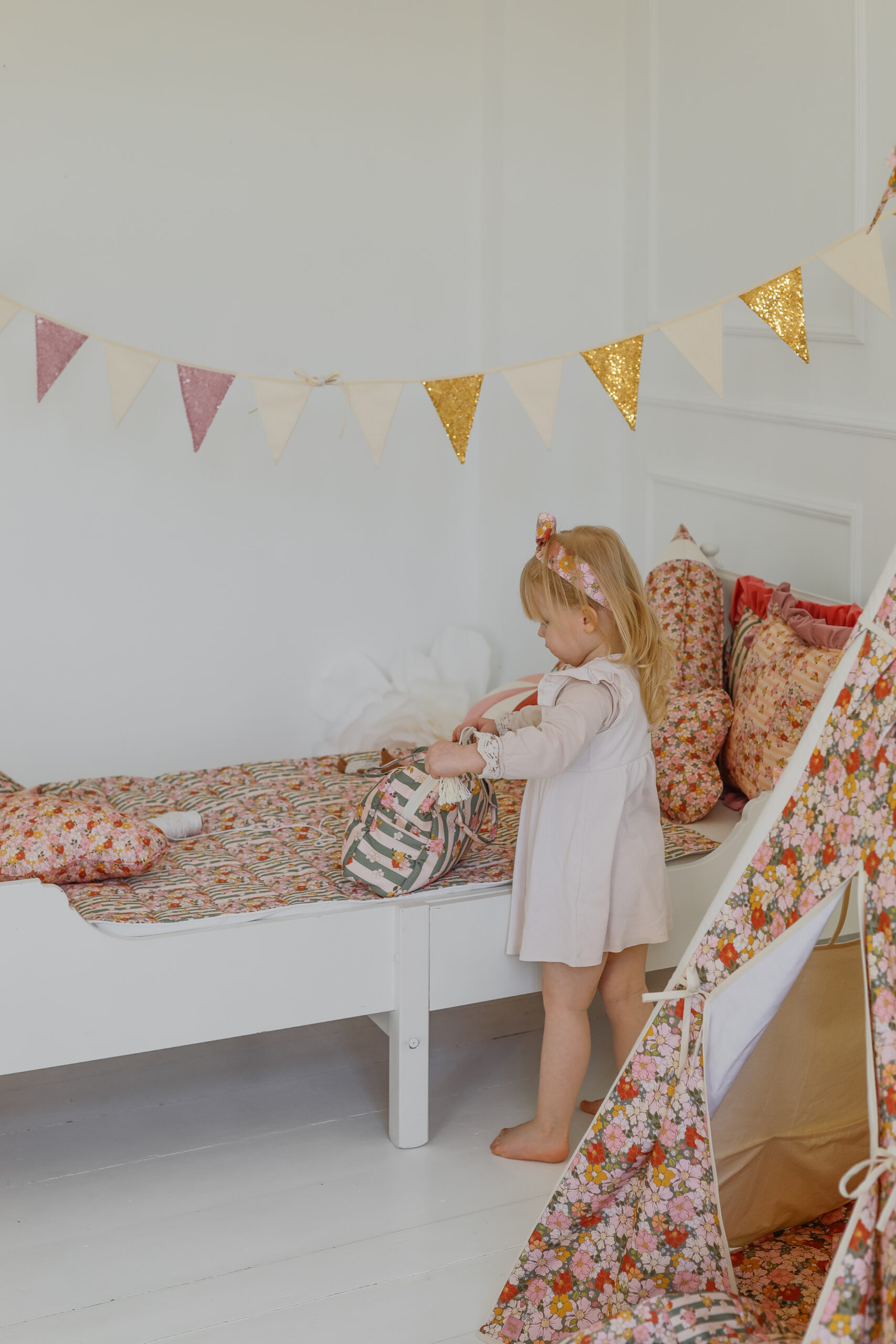 Mała dziewczynka w jasnej sukience stoi przy białym łóżku z kolorową pościelą w pokoju dziecięcym z tipi