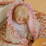 Małe dziecko otulone różowo-kwiatowym kocykiem siedzi obok jasnego drewnianego stolika w przytulnym wnętrzu