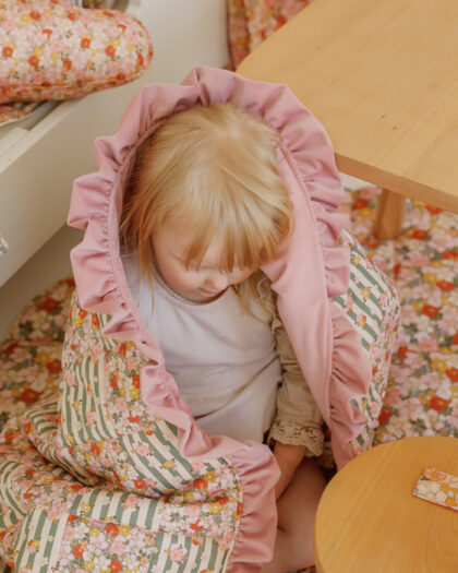 Małe dziecko otulone różowo-kwiatowym kocykiem siedzi obok jasnego drewnianego stolika w przytulnym wnętrzu