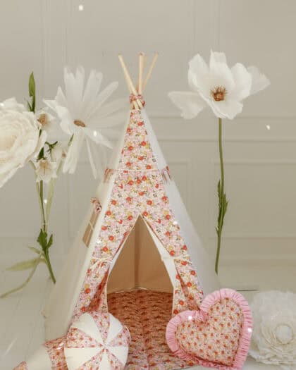 Białe tipi z różowym wzorem kwiatowym, obok trzy poduszki we wzory florystyczne i serce oraz dekoracje z dużych białych kwiatów na jasnym tle i białej podłodze.