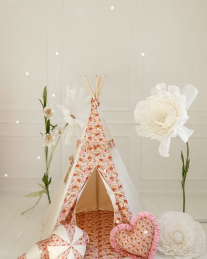 Mały biały pokój dziecięcy z tipi w kwiatowym wzorze w odcieniach różu i pomarańczu, otwartym wejściem, na podłodze miękka mata w tym samym wzorze, obok tipi trzy poduszki: w kształcie serca, stożka i kulki, wokół tipi duże ozdobne białe kwiaty na tle białej ściany z delikatnym reliefem.