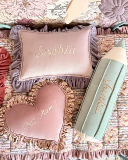 Trzy ozdobne poduszki na łóżku z kwiatową narzutą, poduszki w kształcie prostokąta z napisem Sophia, serca z napisem Best Mum! oraz ołówka z napisem Luna, w pastelowych kolorach różu, fioletu i mięty