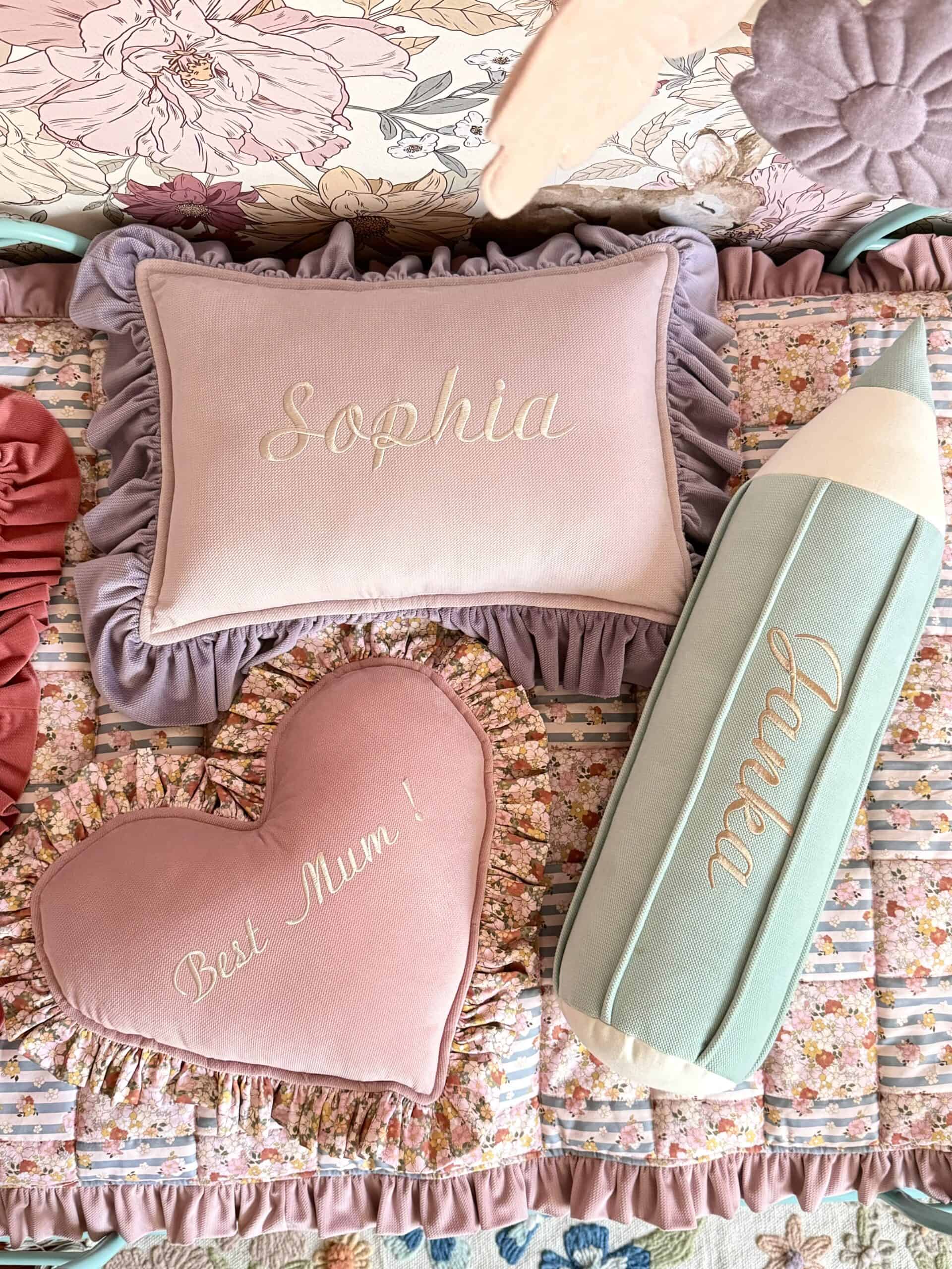 Trzy ozdobne poduszki na łóżku z kwiatową narzutą, poduszki w kształcie prostokąta z napisem Sophia, serca z napisem Best Mum! oraz ołówka z napisem Luna, w pastelowych kolorach różu, fioletu i mięty