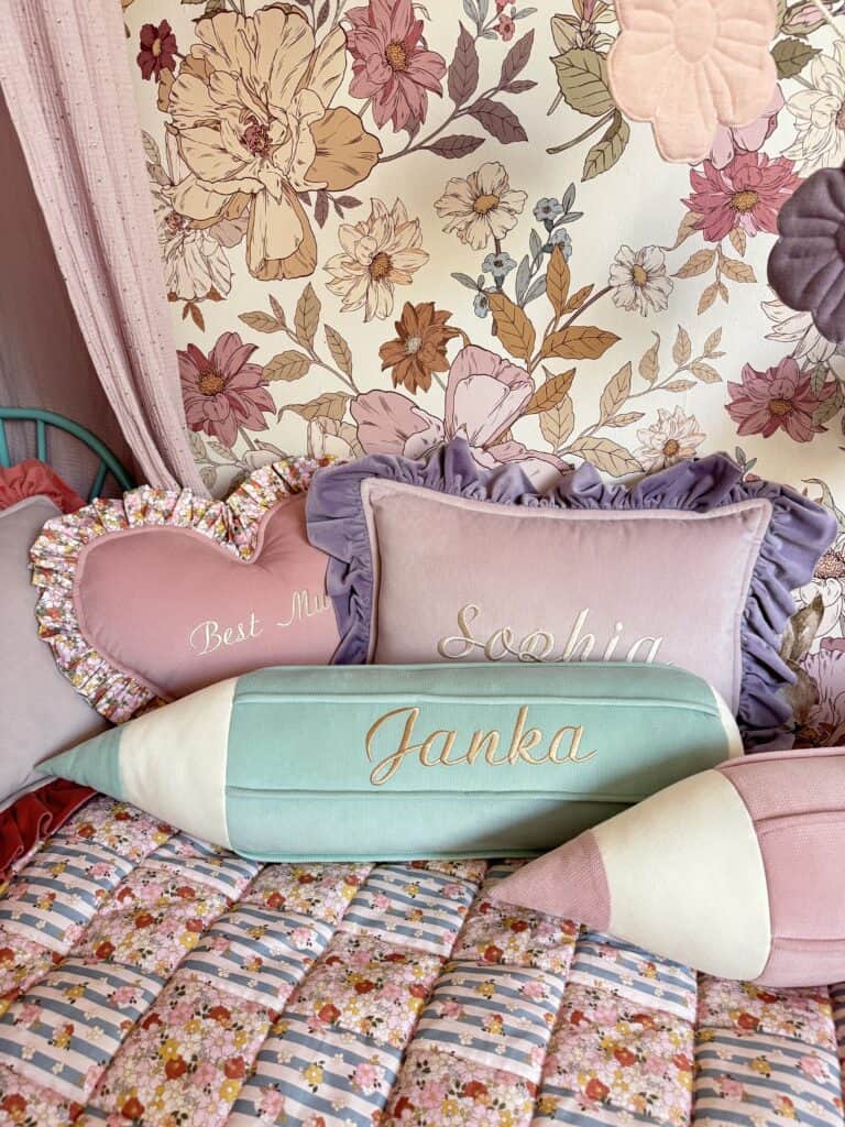 Miękkie poduszki na łóżku z kolorową pościelą, w tym różowa poduszka w kształcie serca z napisem "Best Mum", prostokątna poduszka z falbanką i napisem "Sophia", oraz poduszki w kształcie kredki z imionami "Janka" i różowa na tle ściany z kwiatowym wzorem