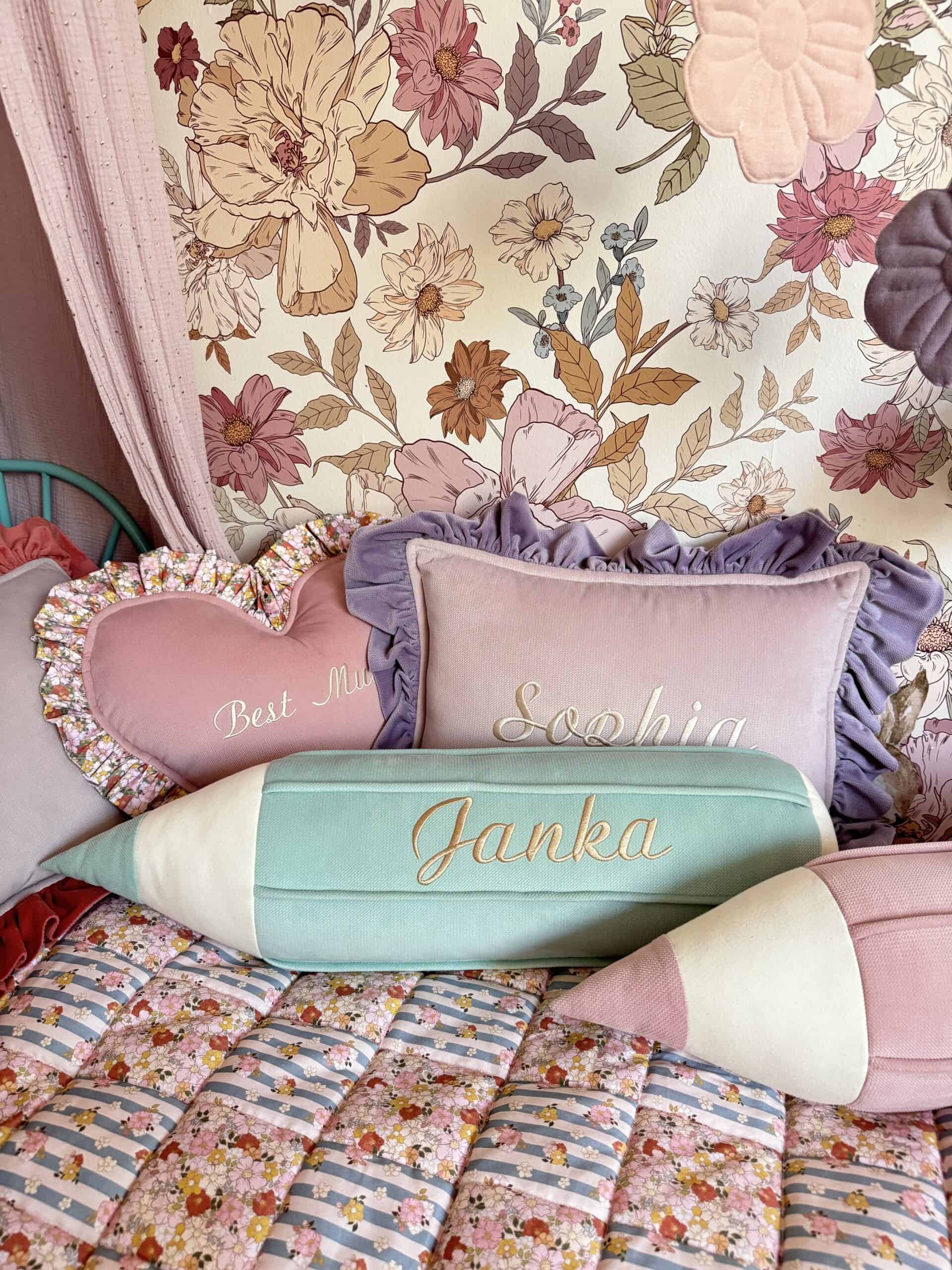 Miękkie poduszki na łóżku z kolorową pościelą, w tym różowa poduszka w kształcie serca z napisem "Best Mum", prostokątna poduszka z falbanką i napisem "Sophia", oraz poduszki w kształcie kredki z imionami "Janka" i różowa na tle ściany z kwiatowym wzorem