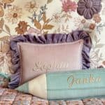 Różowa poduszka z falbanami i haftowanym imieniem Sophia oraz zielona poduszka w kształcie ołówka z imieniem Janka na tle kwiatowej tapety i kolorowej narzuty w kwiaty.