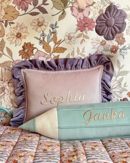 Różowa poduszka z falbanami i haftowanym imieniem Sophia oraz zielona poduszka w kształcie ołówka z imieniem Janka na tle kwiatowej tapety i kolorowej narzuty w kwiaty.