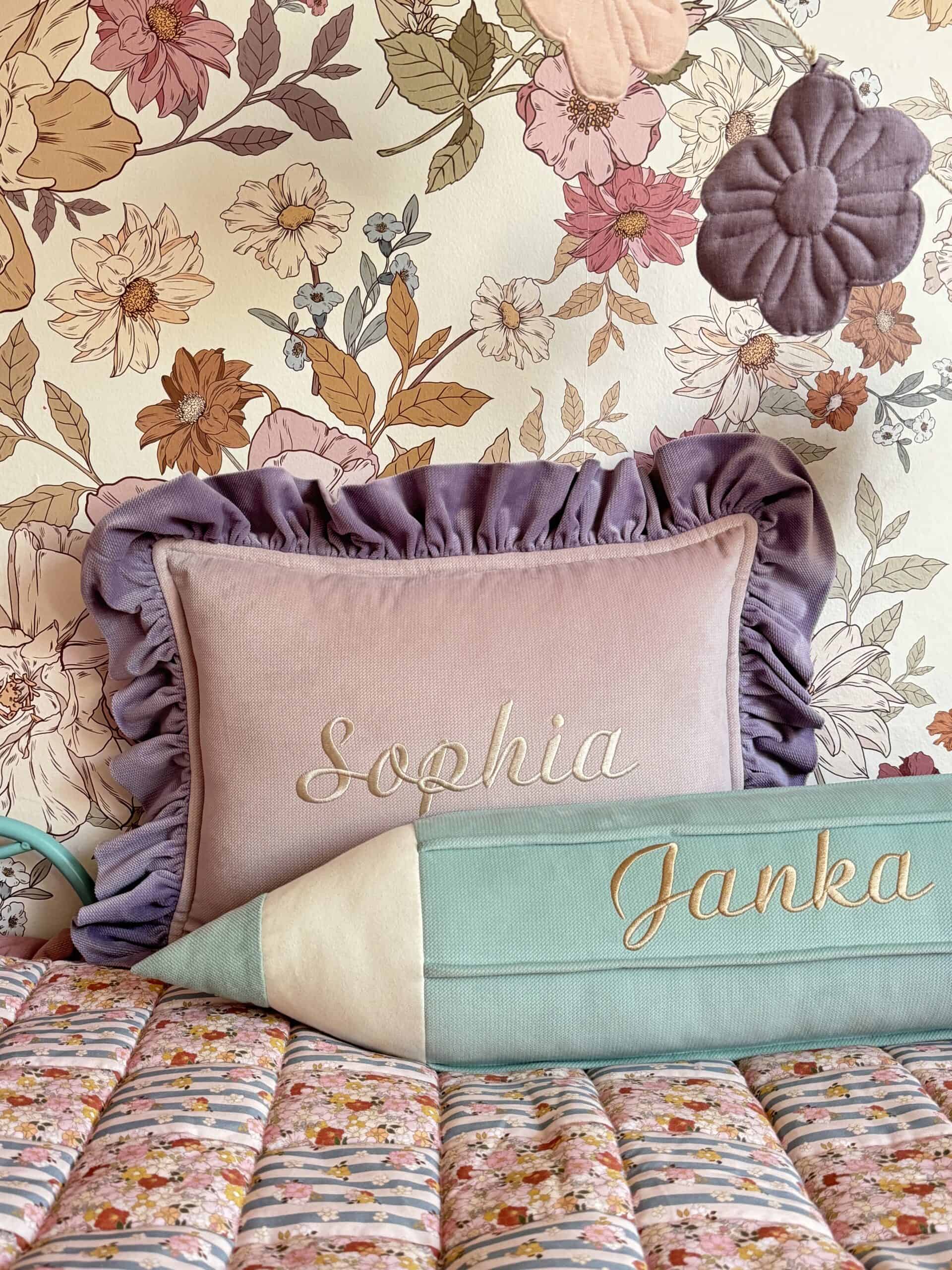 Różowa poduszka z falbanami i haftowanym imieniem Sophia oraz zielona poduszka w kształcie ołówka z imieniem Janka na tle kwiatowej tapety i kolorowej narzuty w kwiaty.
