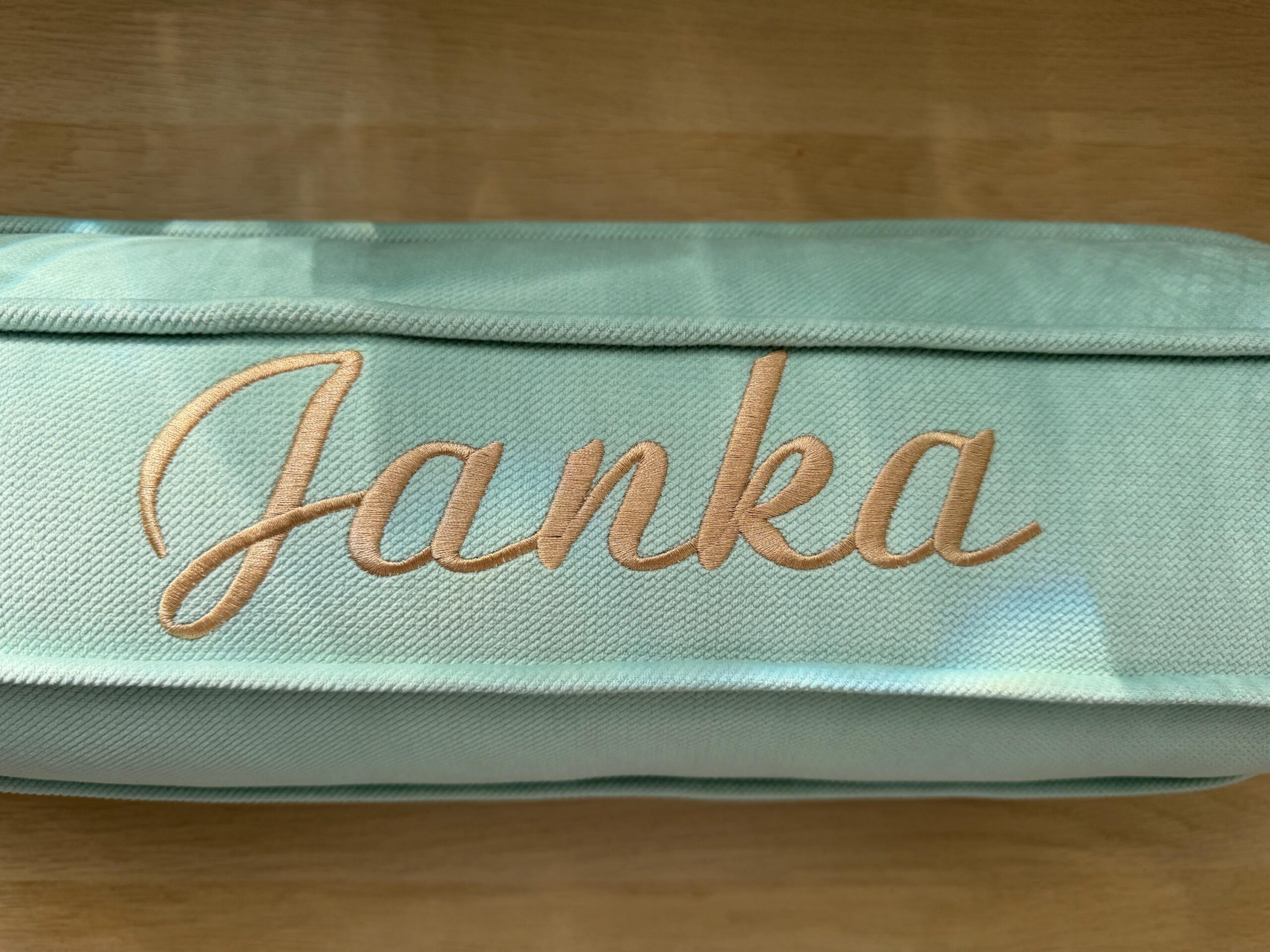 Miętowa tkanina z haftowanym beżowym napisem Janka na jasnym drewnianym tle