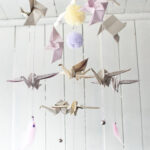Ręcznie wykonany mobil dziecięcy z origami w kształcie ptaków, pomponów i delikatnych piórek, zawieszony na białym, rozgałęzionym kijku przed jasną drewnianą ścianą.
