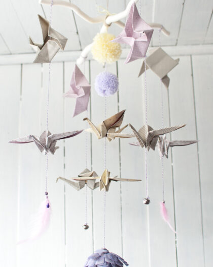 Ręcznie wykonany mobil dziecięcy z origami w kształcie ptaków, pomponów i delikatnych piórek, zawieszony na białym, rozgałęzionym kijku przed jasną drewnianą ścianą.