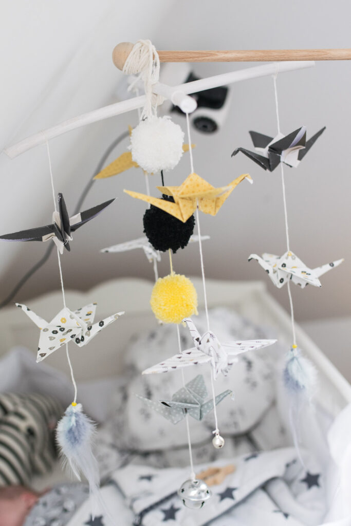 Zbliżenie na ręcznie wykonany biało-czarny i żółty mobil nad łóżeczkiem dziecięcym, zawieszony na drewnianym ramieniu, z origami ptaków, kolorowymi pomponami i piórkami.