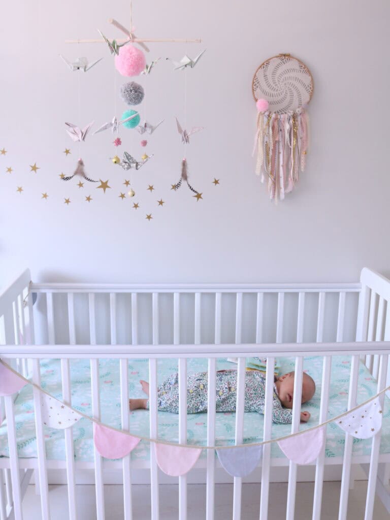 Białe łóżeczko dziecięce z niemowlęciem śpiącym na niebieskiej pościeli z motywem zwierząt, nad łóżeczkiem wiszą dekoracje z origami i pomponami oraz łapacz snów, na poręczy girlanda z różowymi, białymi i niebieskimi chorągiewkami z gwiazdkami na białej ścianie w tle.