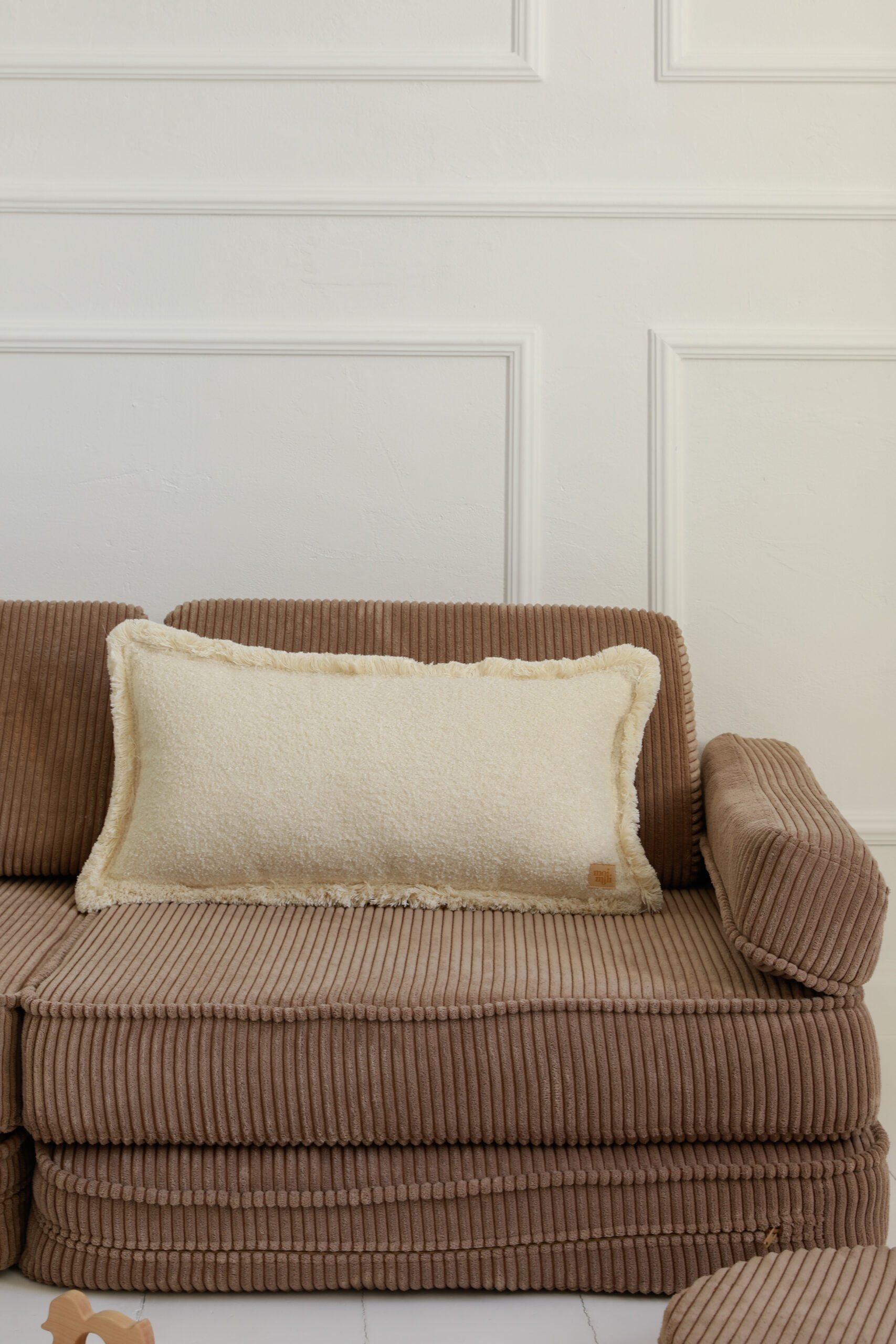 Beżowa poduszka dekoracyjna na brązowej, prążkowanej kanapie w jasnym wnętrzu z białą ścianą