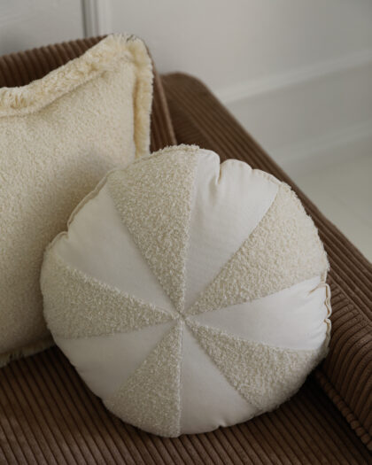 Okrągła jasna poduszka z naprzemiennym wzorem materiałów na brązowej kanapie obok kwadratowej poduszki